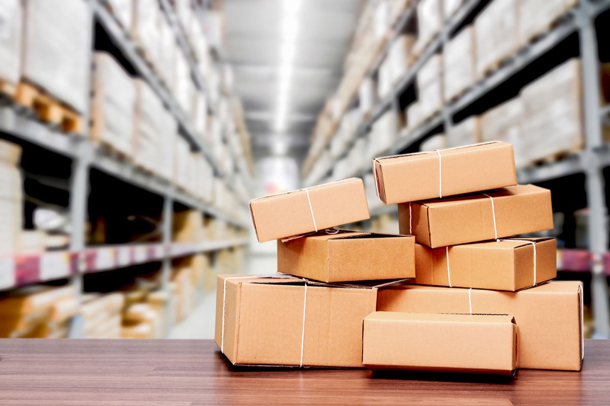 Stack of taped cardboard parcels on a wooden counter in front of blurred warehouse shelving aisles