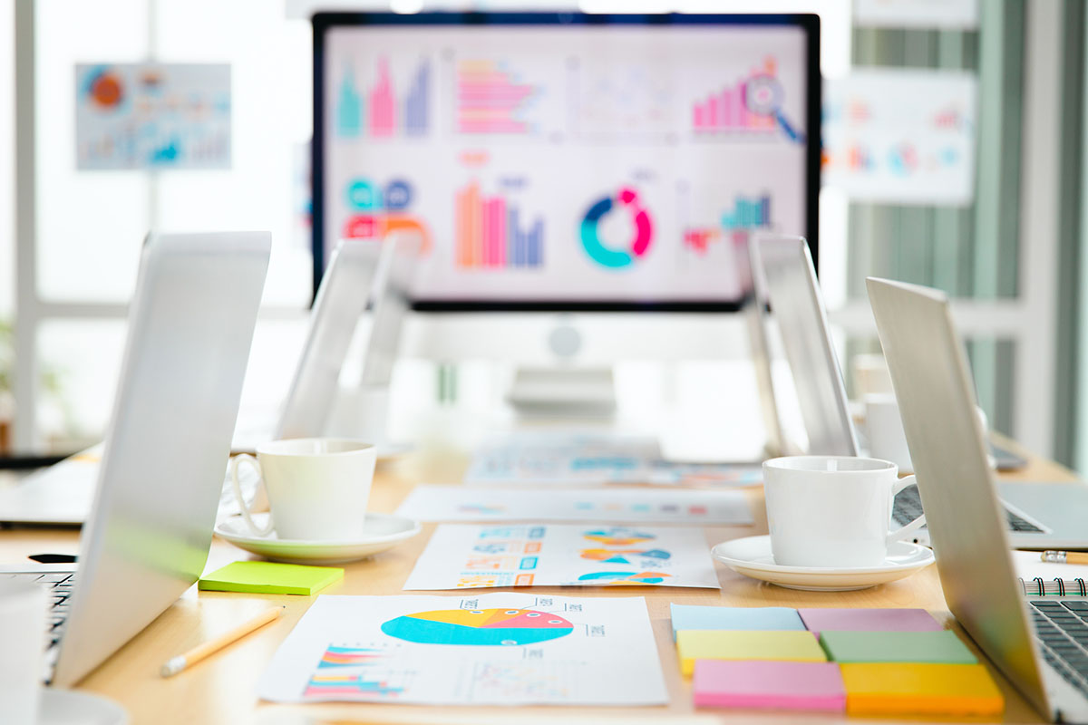 Conference table with laptops, coffee cups, sticky notes and printed charts in front of a monitor showing colorful business analytics in a bright meeting room