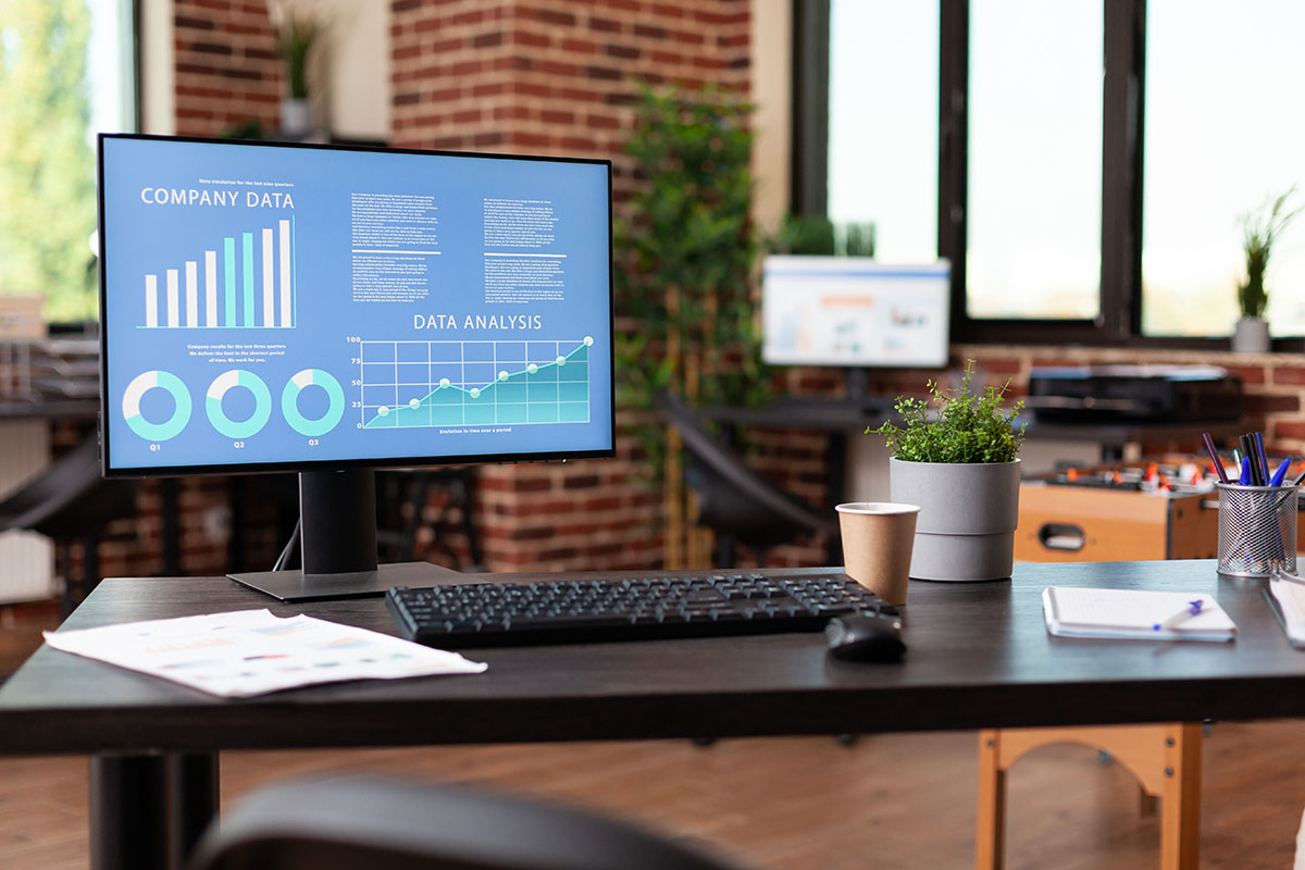 Desktop monitor showing company data bar charts and a data analysis line graph on a desk in an empty modern office with brick walls