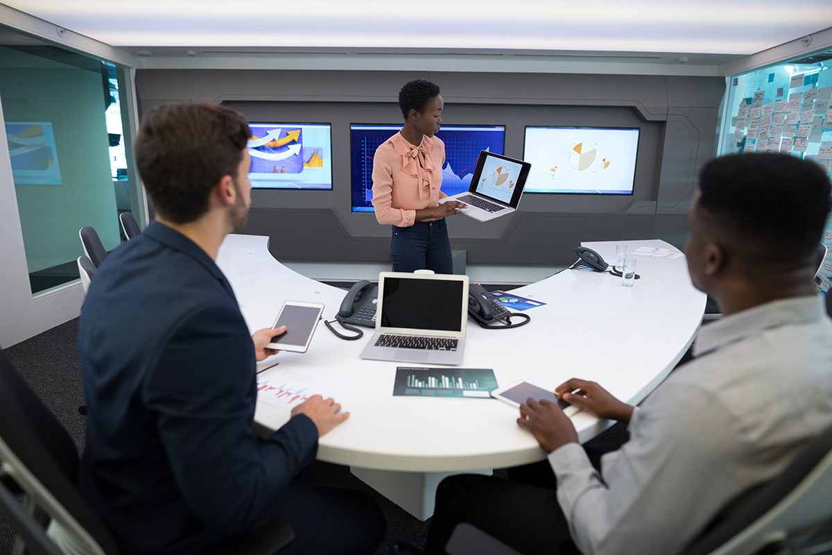 Executive presenting charts on a laptop to two colleagues at a curved white conference table in a high-tech meeting room with wall screens