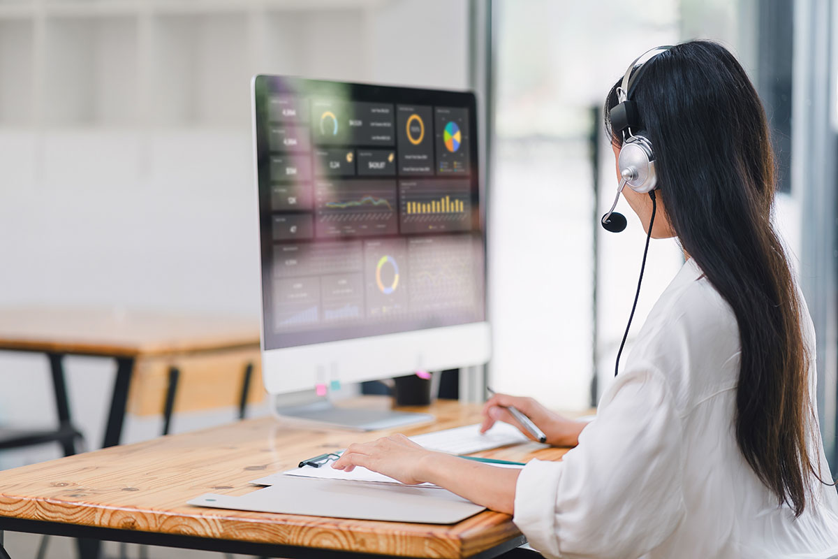 Customer service agent wearing a headset, seen from behind, working at a wooden desk in front of a monitor showing a performance analytics dashboard