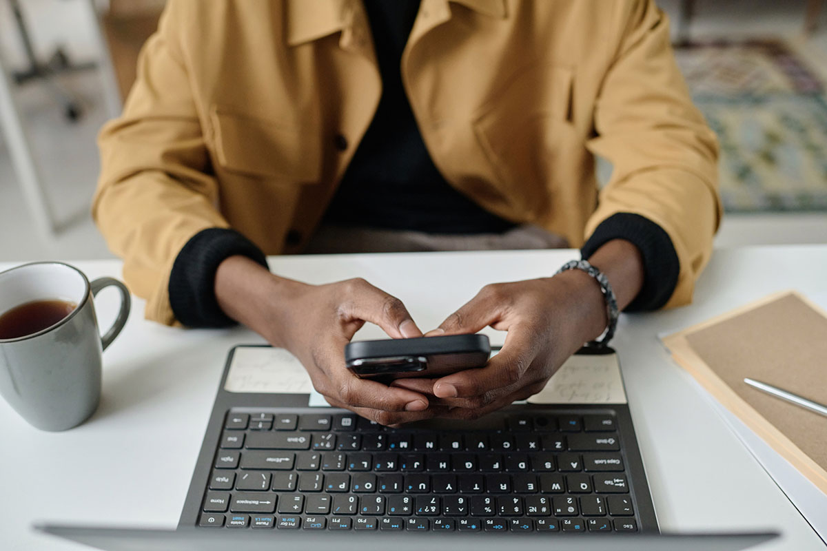 Close-up of an employee&rsquo;s hands typing on a smartphone above an open laptop on a white desk, beside a mug of tea and a folder
