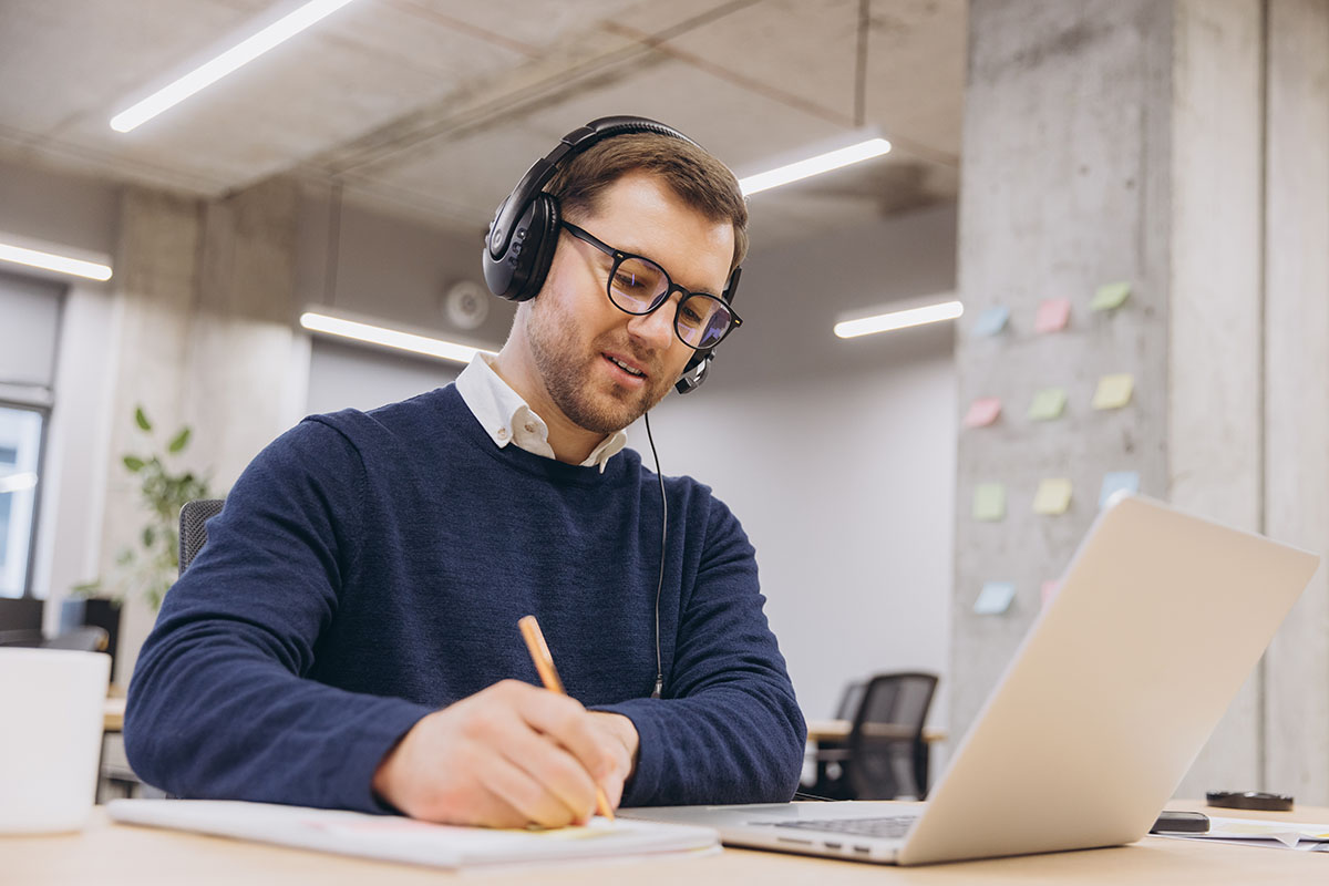 Man wearing headphones and glasses on a video call at a laptop, taking notes at a desk in a largely empty open-plan office