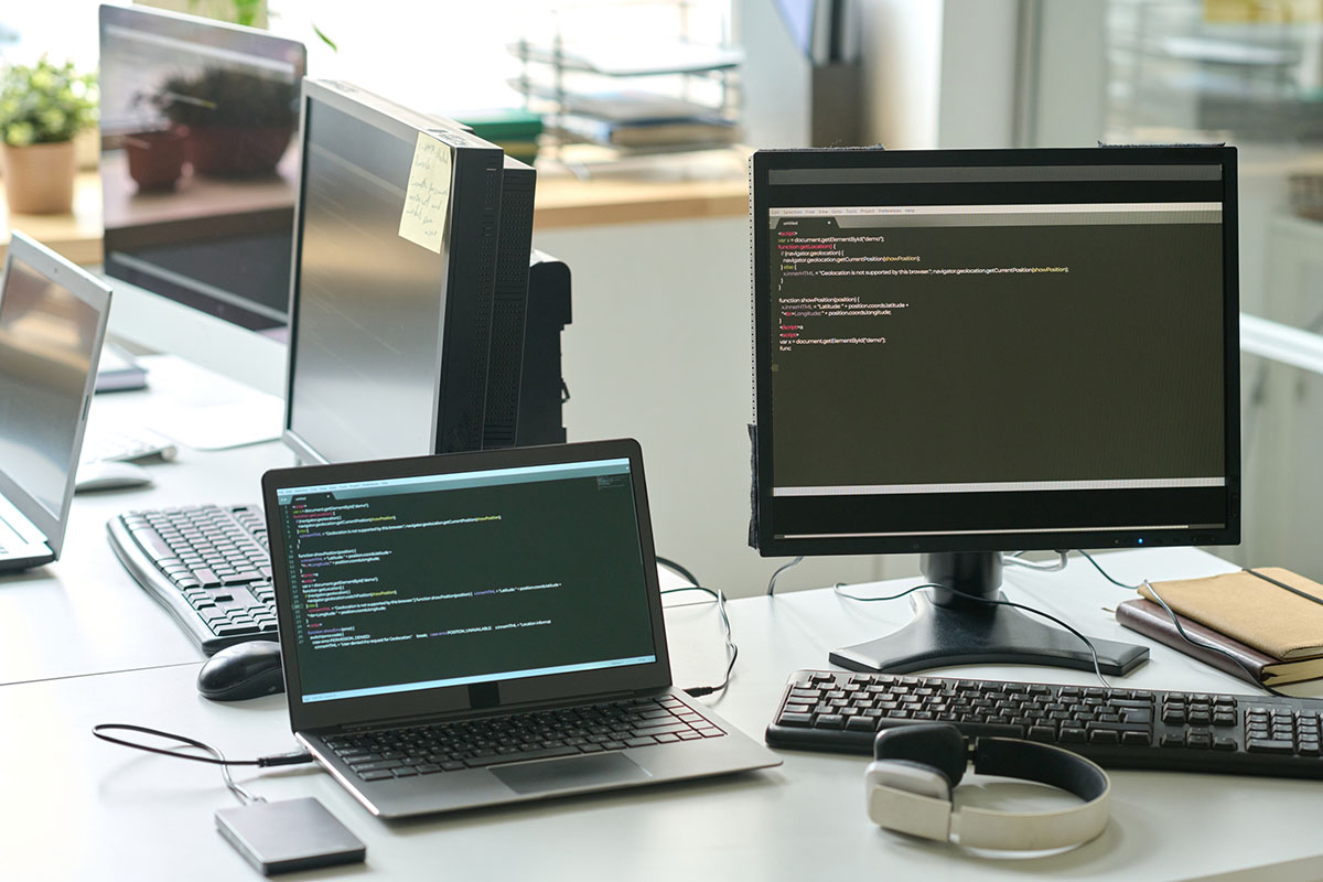Office desk with a laptop and several computer monitors showing software windows, with keyboards, headphones, and notebooks beside them