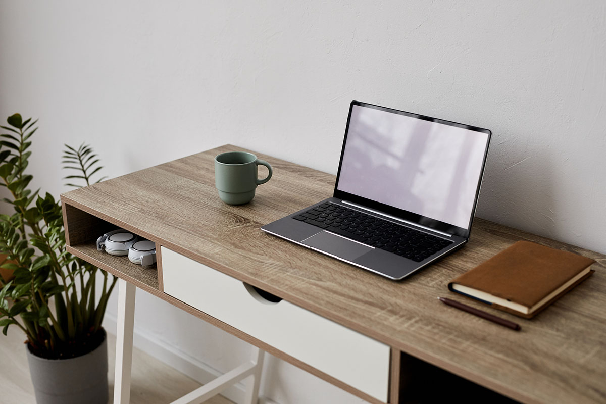 An empty home office desk with an open laptop, a green coffee mug, a leather notebook and pen, and headphones on the shelf beside a potted plant