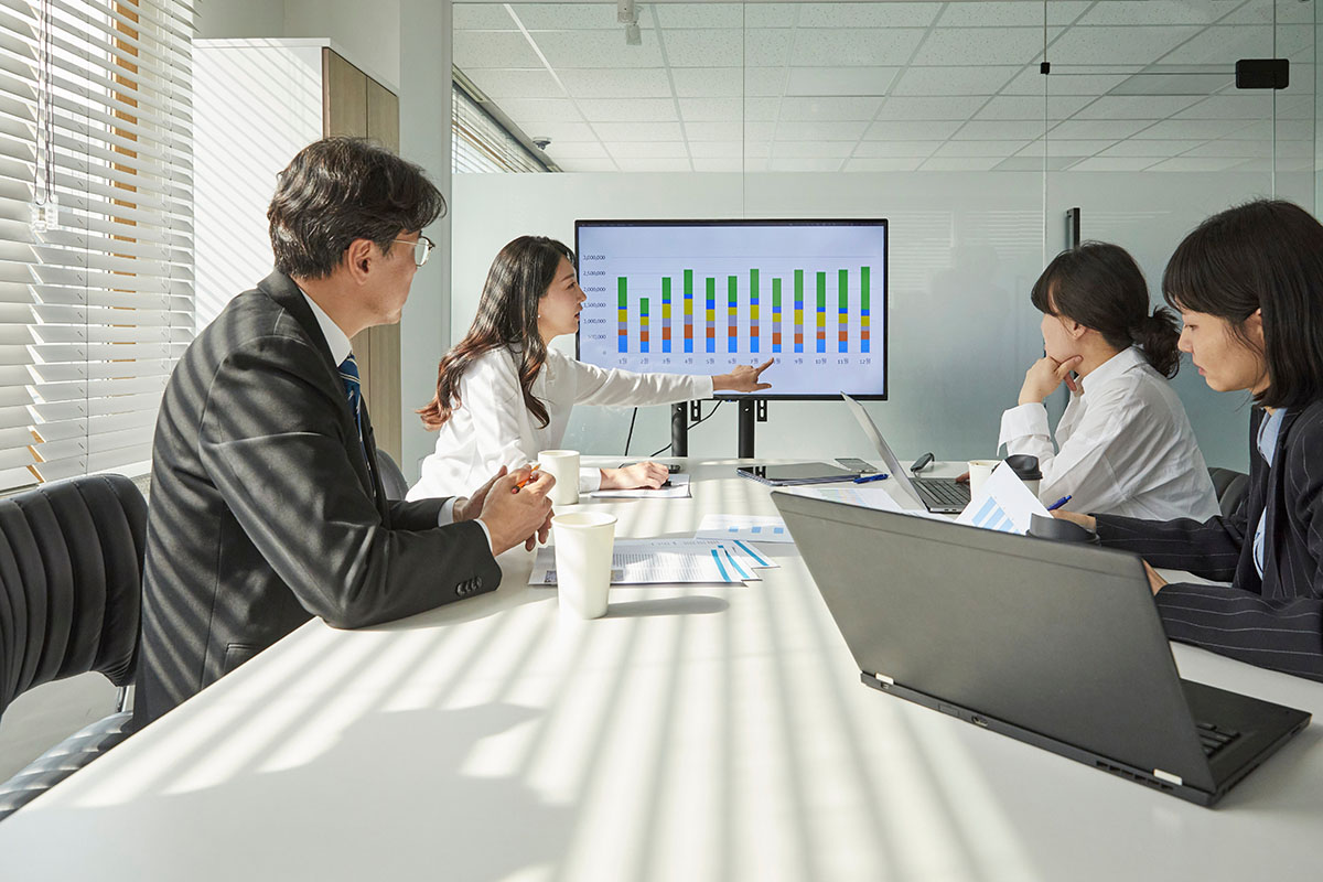 Business team in a sunlit conference room reviewing a bar chart on a large screen as a colleague points to the data