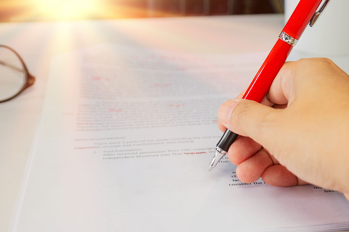 Close-up of a hand marking corrections in red pen on a printed document, with reading glasses resting on the desk beside it